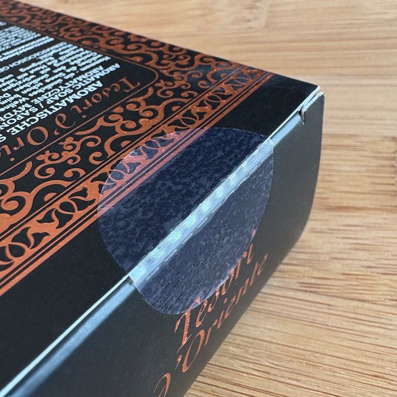Oplossingen Cosmetica: Zwarte verpakking met decoratieve, oranje patronen en tekst. De flap van de verpakking is verzegeld met een transparante sluitsticker.