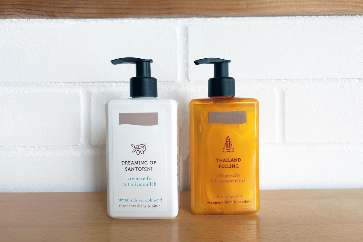 Twee pompflessen met vloeibare zeep: een witte fles met een wit etiket en een oranje fles met een transparant folie-etiket in een no-label stijl, op een houten oppervlak voor een witte bakstenen muur.