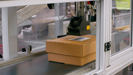 Een doos wordt op een transportband in een geautomatiseerde verpakkingsinstallatie verwerkt. Het geactiveerde InNo-Liner etiket hecht onmiddellijk op absorberende oppervlakken en bereikt binnen korte tijd zijn volledige kleefkracht.