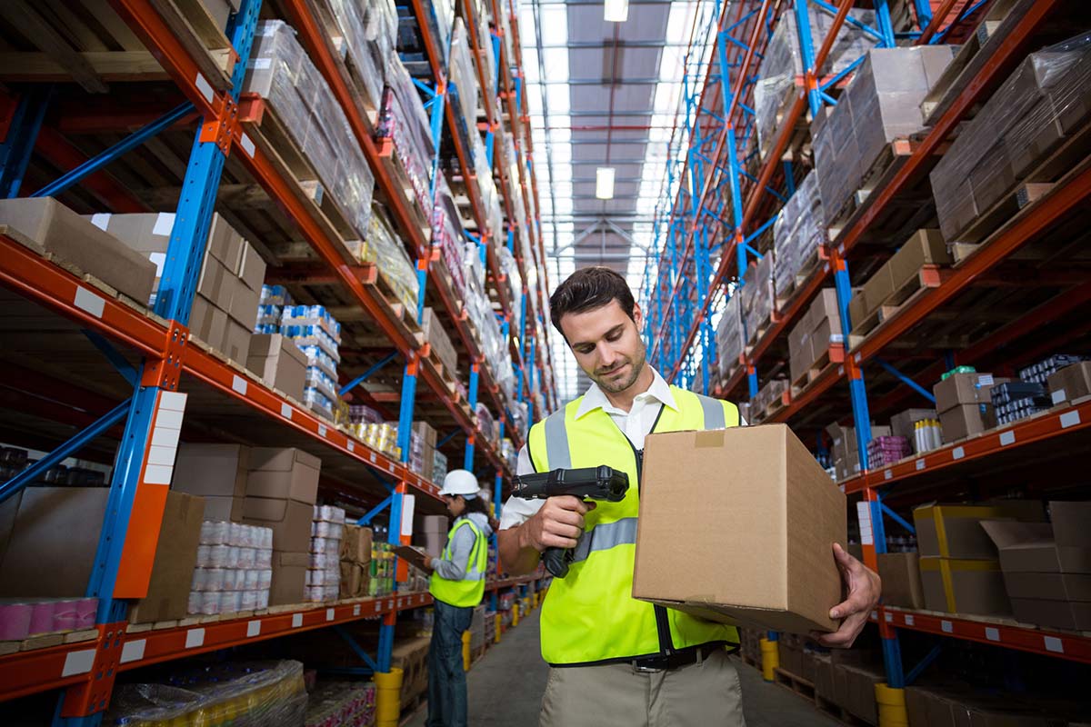 Verzend- en palletlabels: Magazijnmedewerker scant een transportdoos, met een hoogbouwmagazijn op de achtergrond.
