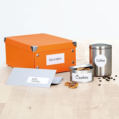 Bedrukte universele etiketten op een oranje doos, zilveren koffiecontainer, blik met koekjes en een grijze envelop op een houten tafel.