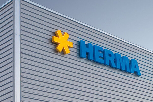 Gevel van een modern gebouw met het logo 'HERMA' in blauwe letters en een geel symbool.