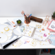 Bürotisch mit Schreibutensilien, Notizbuch, Aufklebern, Pflanzen und verschiedenen Bastelmaterialien.
