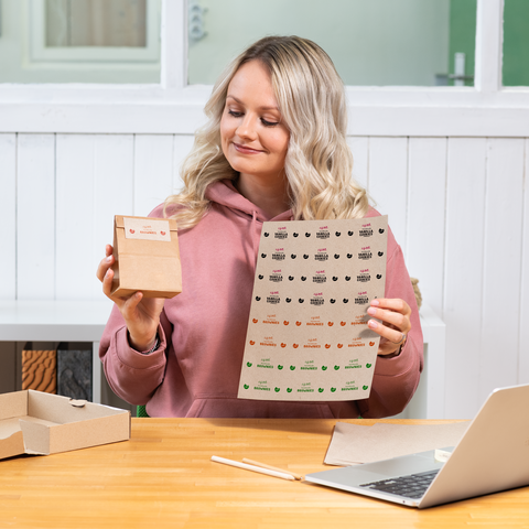 Eine Frau mit blonden, lockigen Haaren hält eine kleine Verpackung und ein Blatt mit Aufklebern in einer hellen Umgebung.