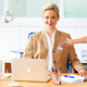 Een vrouw met blond haar zit aan een bureau, draagt een blauwe blazer en glimlacht. Op de tafel staat een laptop en schrijfmateriaal.