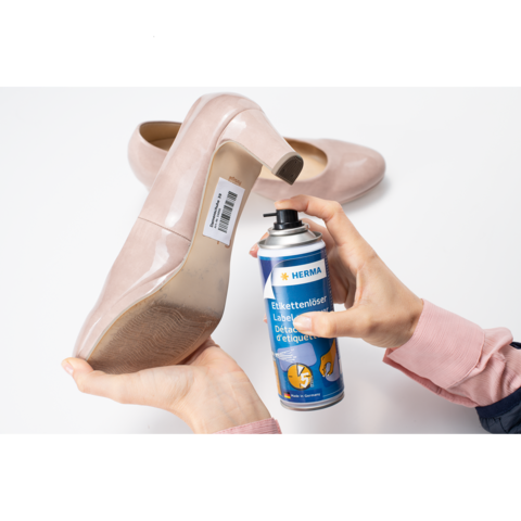 Eine Hand hält einen beigen High Heel mit einem Etikett, während die andere Hand eine Spraydose mit Etikettenentferner hält.