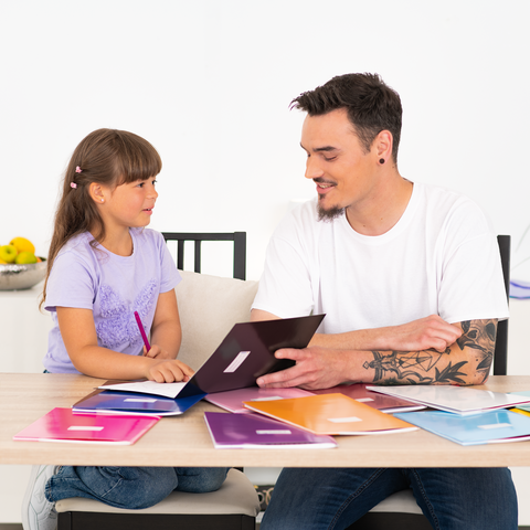 Ein Mann und ein Mädchen sitzen an einem Tisch, umgeben von bunten Heften. Sie schauen sich ein Heft an und scheinen zu kommunizieren.