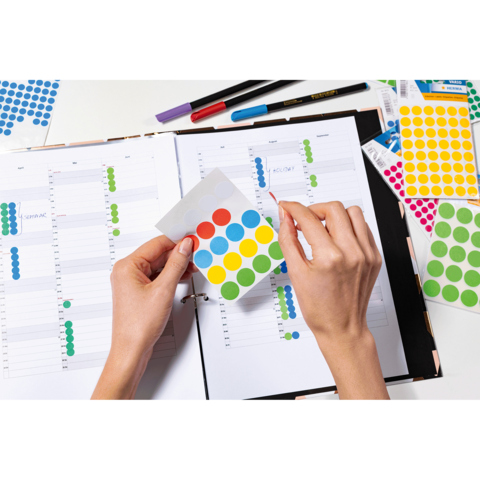 Eine Person hält ein Blatt mit bunten Aufklebern über einem geöffneten Planer, der mit Notizen und weiteren Aufklebern gefüllt ist.