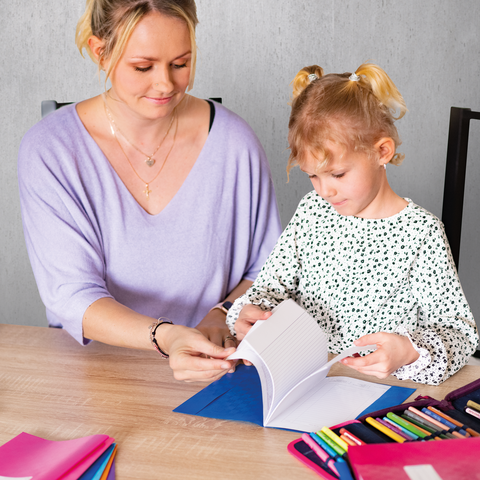 Eine Frau und ein kleines Mädchen sitzen an einem Tisch, während sie gemeinsam in einem Notizbuch blättern. Bunte Stifte liegen verstreut.