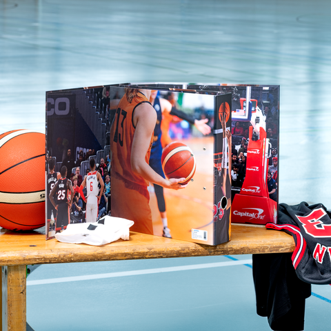 Ein Basketball, ein Trikot und ein T-Shirt liegen auf einem Tisch, umgeben von einer Basketballspiel-Szene auf einer Box.