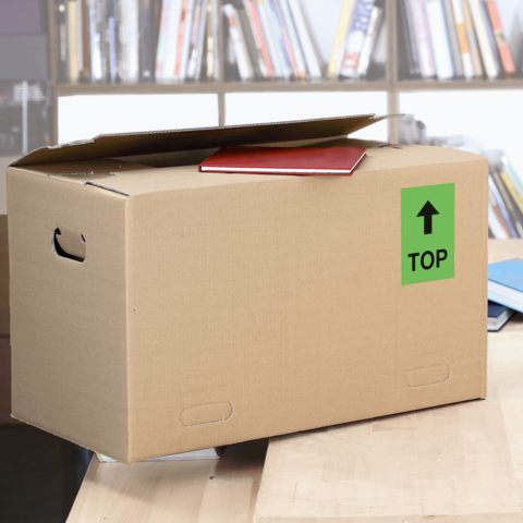 Kartonbox auf einem Tisch, mit einem roten Heft und einem grünen Aufkleber "TOP" auf der Seite. Hintergrund mit Bücherregalen.