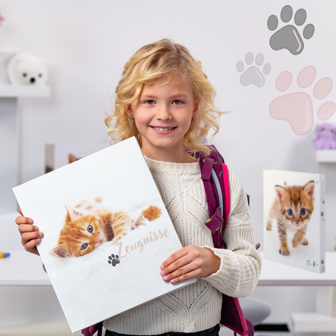 Een meisje met blonde krullen houdt een map met een kattenfoto vast. Op de achtergrond zijn er meer mappen en speelgoed te zien.