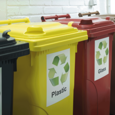 Drei Mülltonnen in verschiedenen Farben: gelb für Plastik, rot für Glas und schwarz für Restmüll, mit Recycling-Symbolen.