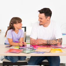 Ein Mann und ein Mädchen sitzen an einem Tisch, umgeben von bunten Farbkarten. Sie scheinen ein kreatives Projekt zu besprechen.