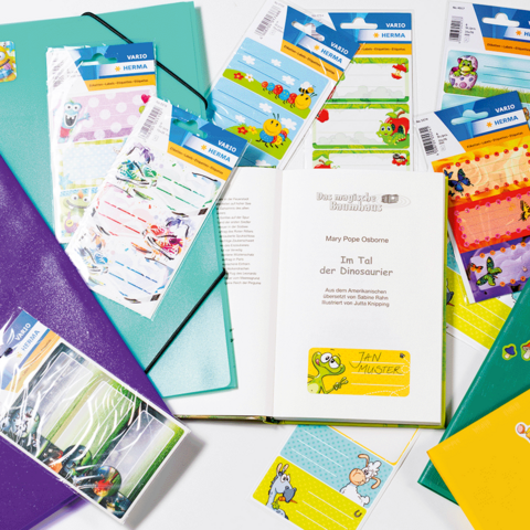 Eine Hand schreibt in ein Notizbuch, während mehrere bunte Aufkleber und ein Buch mit Illustrationen auf dem Tisch liegen.