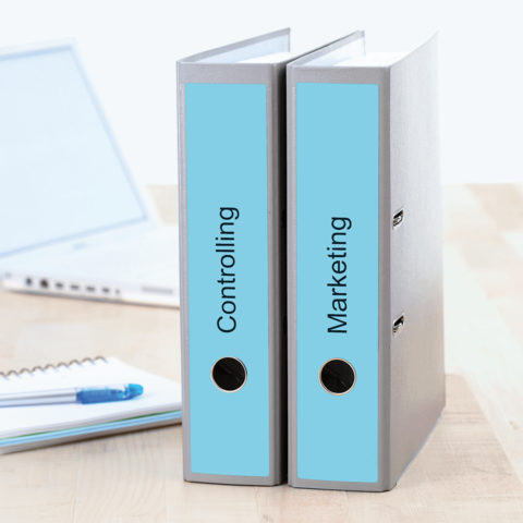 Zwei Aktenordner mit den Beschriftungen "Controlling" und "Marketing" stehen auf einem Tisch, neben einem Laptop und einem Notizblock.