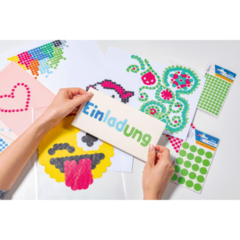 Hände halten eine Einladungskarte mit bunten Aufklebern. Im Hintergrund sind weitere Bastelmaterialien und gestaltete Blätter sichtbar.
