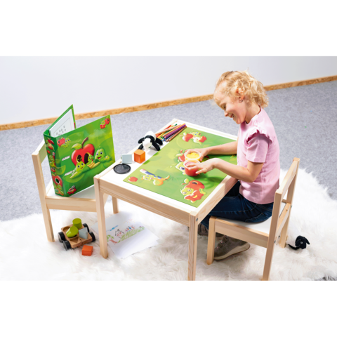 Ein Mädchen sitzt an einem Tisch und gestaltet mit Farben auf einem bunten Spieluntergrund. Neben ihr sind Spielmaterialien und eine Spieleschachtel.