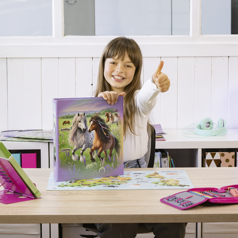 Ein Mädchen sitzt an einem Tisch und zeigt mit dem Daumen nach oben. Vor ihr liegt ein Buch mit Pferdemotiven und ein Puzzle.