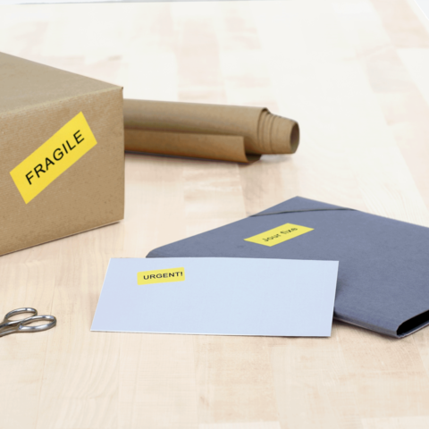 Ein Karton mit dem Aufkleber "FRAGILE" liegt neben einem grauen Umschlag mit dem Aufkleber "DRINGEND!" und einer weißen Karte. Scheren sind ebenfalls sichtbar.