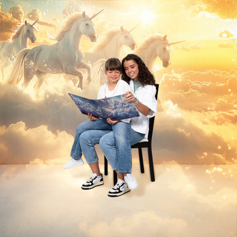 Zwei Mädchen sitzen auf einem Stuhl und betrachten gemeinsam ein Buch mit einem fantasievollen Einhorn-Motiv im Hintergrund.