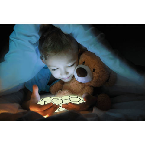 Ein Kind sitzt im Bett, hält einen leuchtenden Gegenstand in den Händen und umarmt einen Teddy. Es ist unter einer Decke.
