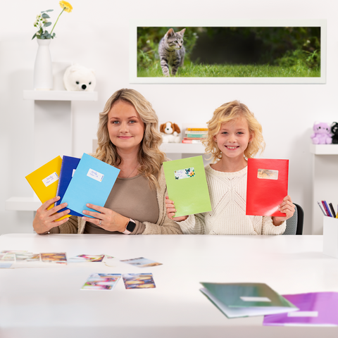 Een vrouw en een meisje houden gekleurde mappen vast terwijl ze aan een tafel met foto's zitten. Op de achtergrond is een kat te zien.