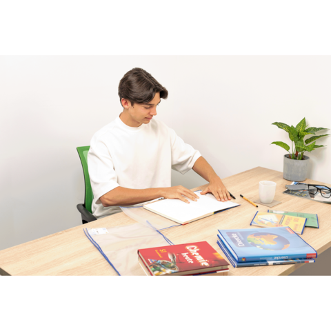 Ein junger Mann sitzt an einem Tisch, blättert in einem Notizbuch und hat Bücher sowie eine Pflanze und eine Tasse auf dem Tisch.