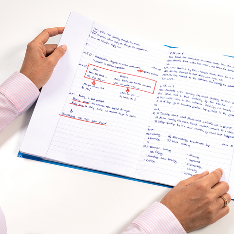 Een persoon houdt een open notitieboek vast met handgeschreven aantekeningen en markeringen in blauwe en rode inkt.