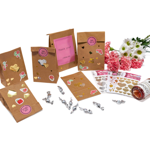 Geschenktüten aus Kraftpapier mit verschiedenen Aufklebern, darunter Herzen und Früchte, umgeben von Blumen und Süßigkeiten.
