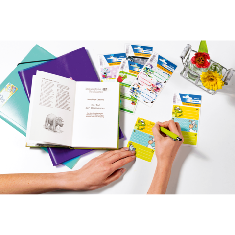 Eine Hand schreibt in ein Notizbuch, während ein offenes Buch und mehrere Karten auf einem Tisch liegen. Blumen in einer Vase sind ebenfalls sichtbar.