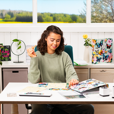 Een vrouw met krullend haar zit aan een bureau, omringd door notities en een laptop, terwijl ze met één hand een gebaar maakt.