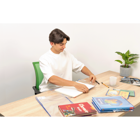 Ein junger Mann sitzt an einem Tisch, blättert in einem Notizbuch und hat Bücher sowie Schreibutensilien vor sich. Eine Pflanze steht im Hintergrund.