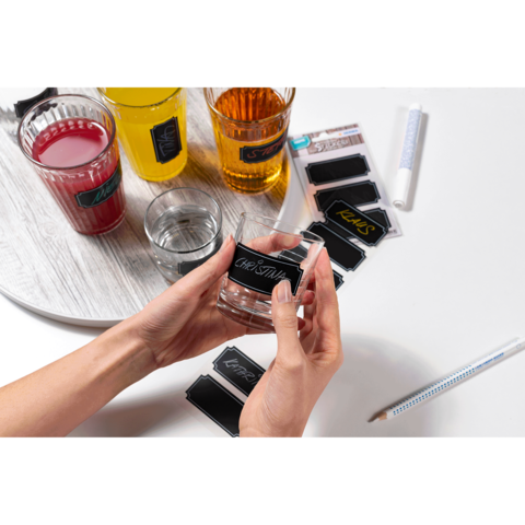 Hände halten ein Glas mit einem Etikett, umgeben von verschiedenen Getränken in Gläsern und Etiketten auf einem Tisch.