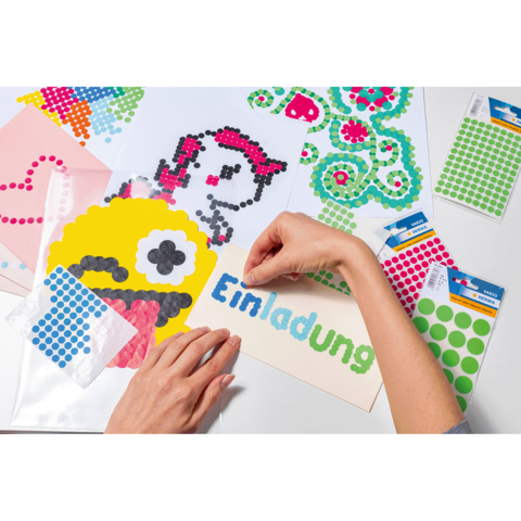 Bunte Bastelmaterialien liegen auf einem Tisch, während eine Hand einen Aufkleber auf eine Einladung platziert.