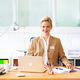 Een vrouw met een blond kapsel zit aan een bureau met een laptop, notitieboek en schrijfgerei, terwijl ze glimlachend in de camera kijkt.