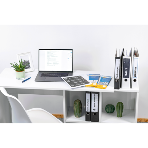 Schreibtisch mit Laptop, Aktenordnern und Unterlagen. Eine Pflanze und ein Bilderrahmen sind ebenfalls sichtbar.