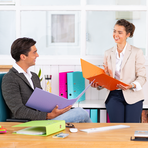 Zwei Personen sitzen an einem Tisch und besprechen Dokumente. Eine Person hält orangefarbene Ordner, während die andere einen lila Ordner in der Hand hat.