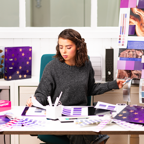 Een persoon zit aan een tafel, omringd door kleurstalen en ontwerpmaterialen, terwijl hij aan een creatief project werkt.