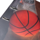Ein Basketball mit sichtbaren Texturen, gehalten von einer Hand, vor einem verschwommenen Basketballkorb im Hintergrund.