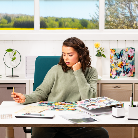 Een jonge vrouw zit aan een bureau, omringd door kleurrijke materialen en notities, terwijl ze nadenkend een pen vasthoudt.