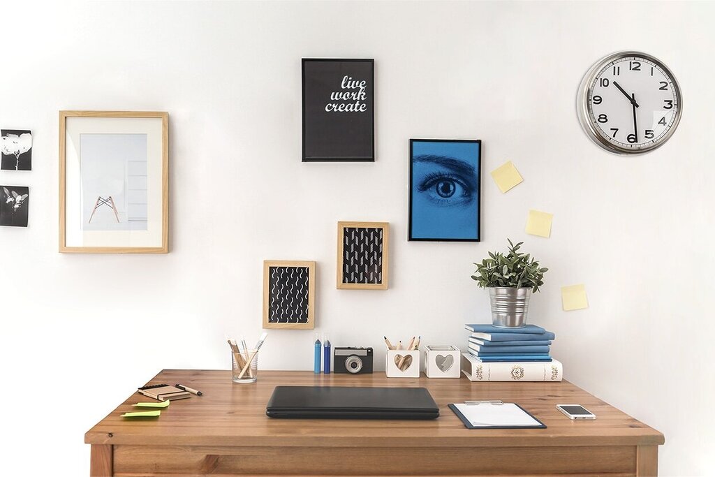 Een bureau met een laptop, decoratieve afbeeldingen aan de muur, een plant en een vloerlamp. Een wandklok geeft de tijd aan.