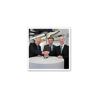 Drie mannen in pakken staan om een tafel en leggen hun handen op een object in het midden.