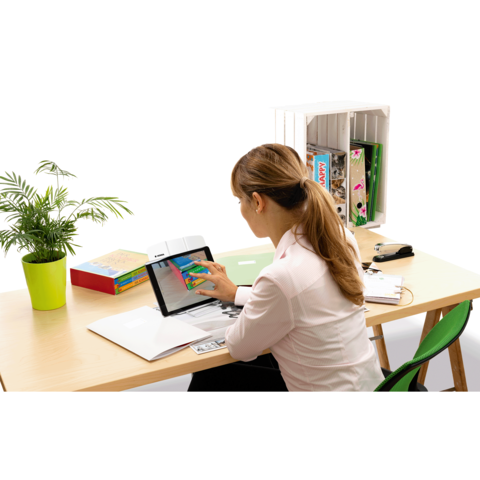 Eine Frau sitzt an einem Schreibtisch und nutzt ein Tablet, während sie auf den Bildschirm tippt. Im Hintergrund sind Bücher und eine Pflanze sichtbar.