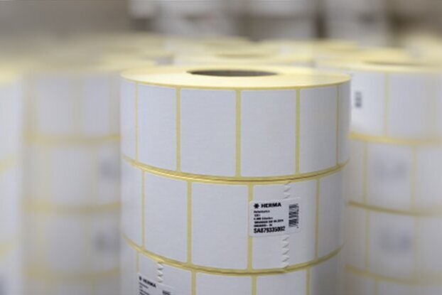 Rollen met witte etiketten, gestapeld in een magazijnomgeving. De etiketten zijn gemarkeerd met gele lijnen.