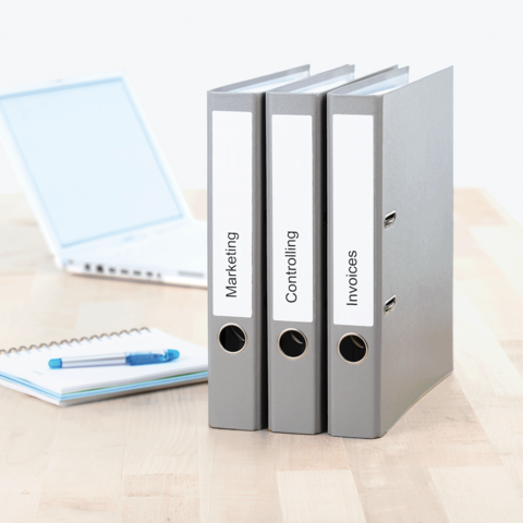Drei graue Ordner mit Beschriftungen „Marketing“, „Controlling“ und „Rechnungen“ stehen auf einem Tisch neben einem Laptop und einem Notizblock.