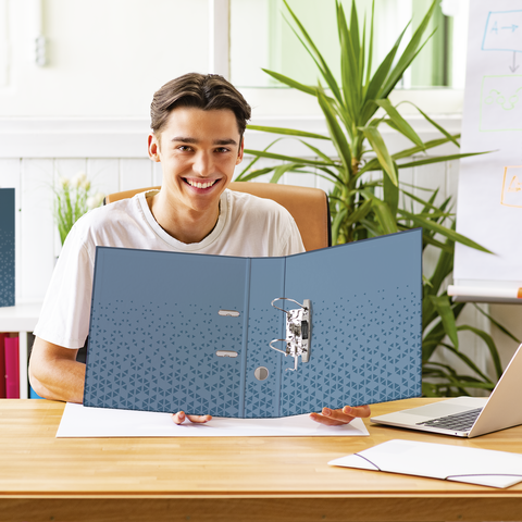 Ein junger Mann sitzt an einem Tisch und hält einen blauen Ordner. Im Hintergrund sind Pflanzen und ein Whiteboard sichtbar.