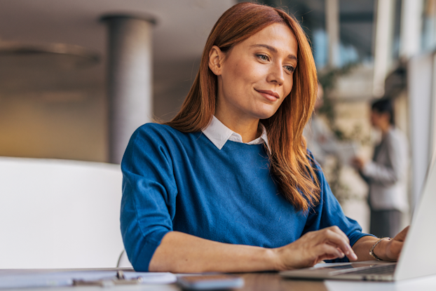 Een vrouw met lange rode haren zit aan een tafel en werkt geconcentreerd aan een laptop in een moderne ruimte.