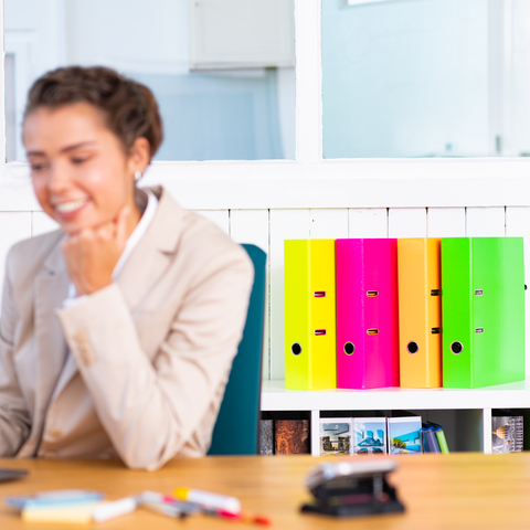 Eine Frau in einem blauen Blazer sitzt an einem Tisch und schaut auf ein Laptop, während bunte Ordner im Hintergrund stehen.