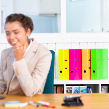 Eine Frau in einem blauen Blazer sitzt an einem Tisch und schaut auf ein Laptop, während bunte Ordner im Hintergrund stehen.